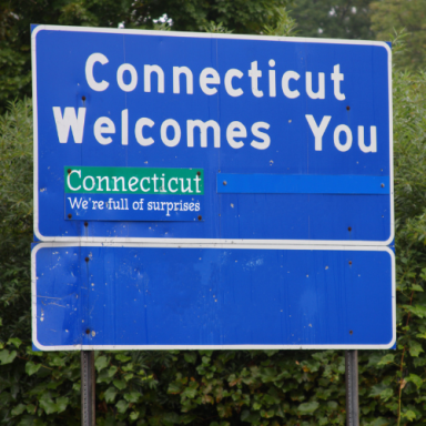 Blue roadside sign welcoming travelers to Connecticut, with "We're full of surprises" text.