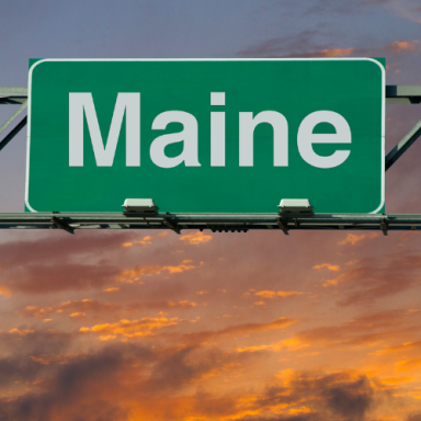 Green road sign displaying "Maine" against a colorful sunset sky.