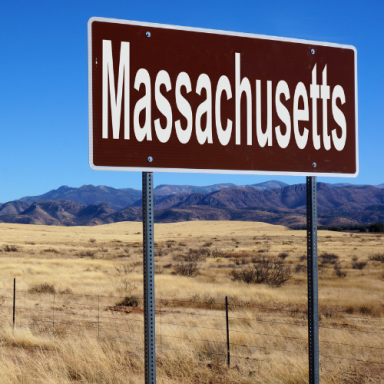 Brown sign with white text reading "Massachusetts" against a clear blue sky and dry landscape.